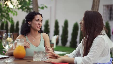 People,-communication-and-friendship-concept---two-women-drinking-orange-juice,-talking-at-outdoor-cafe,-laughing