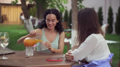 People,-communication-and-friendship-concept---smiling-young-women-drinking-orange-juice-and-talking-at-outdoor-cafe,-smiling
