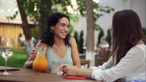 Gente,-Comunicación-Y-Concepto-De-Amistad---Mujeres-Jóvenes-Sonrientes-Bebiendo-Jugo-De-Naranja-Y-Hablando-En-Un-Café-Al-Aire-Libre-En-Verano