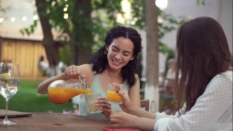 Gente,-Comunicación-Y-Concepto-De-Amistad---Jóvenes-Europeas-Sonrientes-Bebiendo-Jugo-De-Naranja-Y-Hablando-En-Un-Café-Al-Aire-Libre