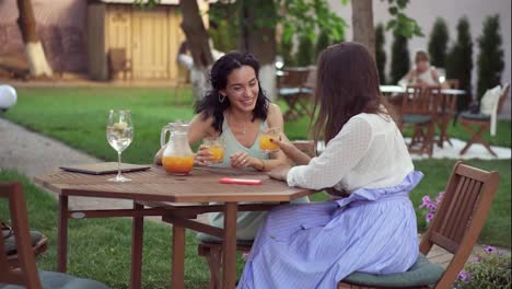 People,-communication-and-friendship-concept---smiling-young-european-women-drinking-orange-juice-at-outdoor-cafe,-clicking