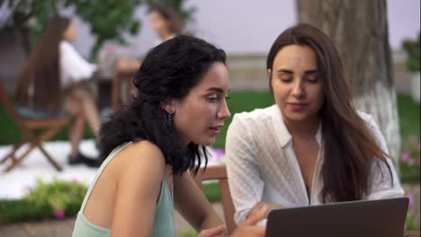 Two-women-sitting-at-the-outdoors-cafe---talking,-sharing-news,-brunette-girl-showing-something-on-laptop-screen,-gesturing