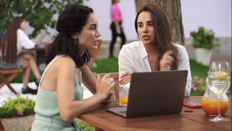 Two-women-sitting-at-the-outdoors-cafe---talking,-sharing-ideas,-showing-something-on-laptop-screen,-gesturing,-arguing