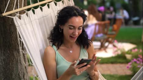 A-girl-with-curly-hair-sits-in-a-hammock-outdoors,-shows-her-smartphone,-clicks-on-the-screen