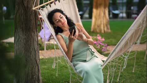 Brunette-woman-relaxing-in-a-hammock-outdoors,-surfing-internet-in-her-smartphone,-clicks-on-the-screen