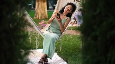 Brunette-woman-relaxing-in-a-hammock-outdoors,-surfing-internet-in-her-smartphone,-clicks-on-the-screen-and-smiling
