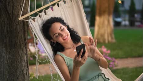 Portrait-Of-a-woman-relaxing-in-a-hammock-outdoors,-surfing-internet-in-her-smartphone,-clicks-on-the-screen-and-smiling