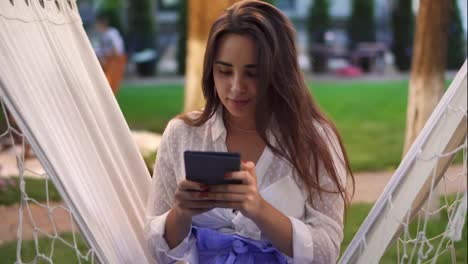 Portrait-of-a-woman-sitting-in-a-hammock-outdoors,-surfing-internet-on-tablet,-clicks-on-the-screen
