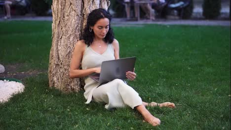 Woman-in-dress-sits-below-tree-at-the-park-on-grass,-holding-laptop-on-her-knees,-typing,-doing-her-work
