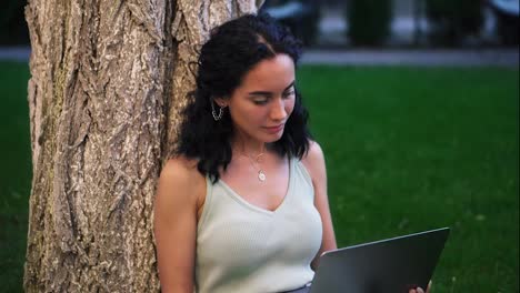 Portrait-of-a-woman-in-dress-sits-below-tree-at-the-park-on-grass,-holding-laptop-on-her-knees,-typing,-doing-her-work