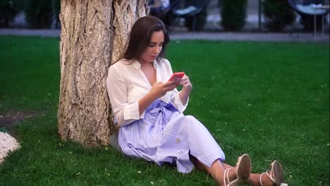 Elegant-woman-sitting-under-a-tree-in-the-park-uses-the-phone-and-smiling