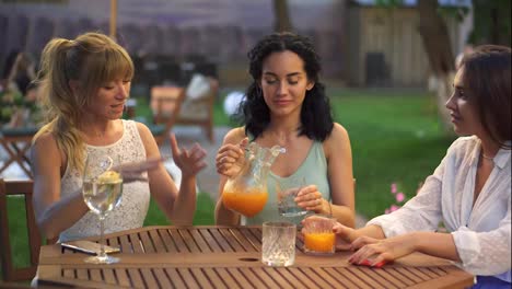 Three-female-friends-sit-in-the-outdoors-cafe,-drink-juice-and-have-fun-communicating,-clinking-with-glasses