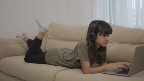 A-teen-girl-uses-a-laptop-while-lying-on-a-couch-in-headphones