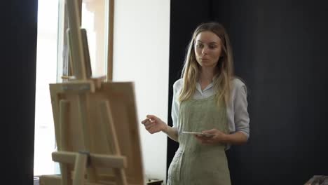 Young-woman-artist-in-apron-painting-picture-on-canvas-in-art-studio