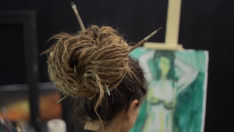 Rear-view-of-female-painter-with-dreadlocks-fixed-by-brushes-painting-on-canvas-at-easel