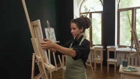 Young-woman-wearing-apron-install-easel-in-workshop
