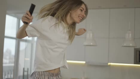 Funny-playful-blonde-woman-dancing-and-singing-at-home-having-fun,-use-remote-controller-as-a-microphone