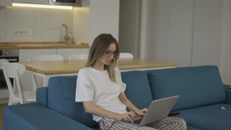 Blonde-woman-in-pajama-sit-on-the-couch-and-typing-on-silver-laptop