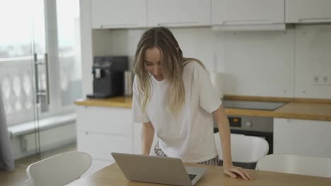 Blonde-Frau-Führt-Online-Videoanrufe-Auf-Dem-Laptop,-Während-Sie-Im-Pyjama-An-Der-Küche-Vorbeigeht