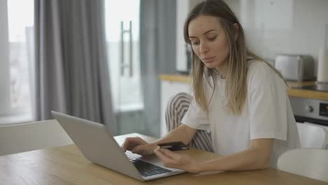 Pretty-girl-is-making-online-payment-holding-bank-card-using-modern-laptop-at-home