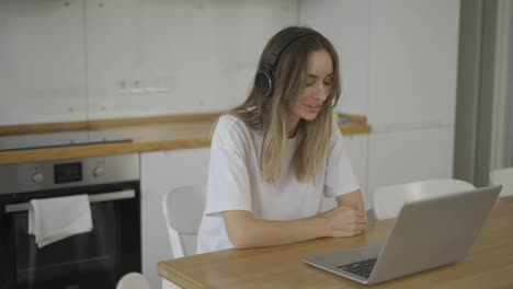 Blonde-woman-make-video-call-online-using-laptop-and-headphones-at-kitchen