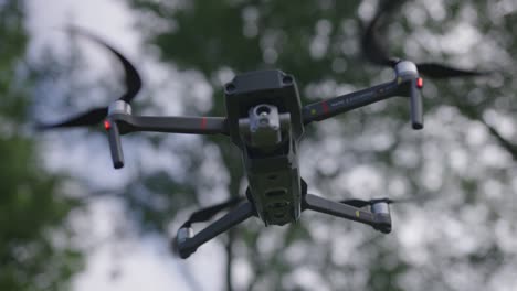 Close-up-shot-of-a-drone-hovering-as-seen-from-low-angle,-pans-slightly-right-to-left-with-trees-in-the-background