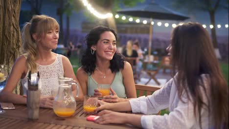 Three-female-friends-sit-in-the-outdoors-cafe,-drink-juice-and-have-fun-communicating,-laughing-in-the-evening