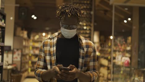 African-American-man-in-mask-busy-with-smartphone-walks-at-the-store