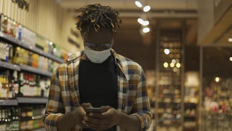 Afroamerikanischer-Mann-In-Maske,-Der-Mit-Smartphone-Beschäftigt-Ist,-Geht-Im-Laden-Spazieren