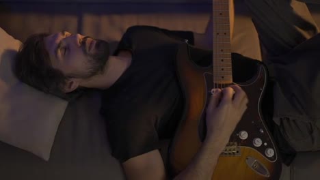 Man-playing-on-guitar-while-lying-on-couch-with-closed-eyes