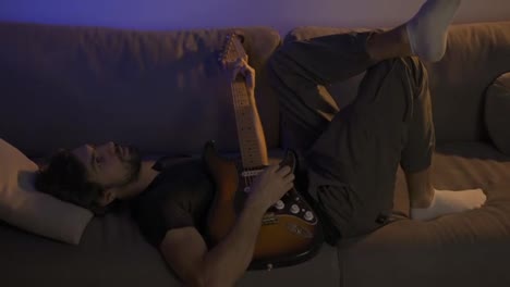Man-composing-music-on-guitar-while-lying-on-sofa