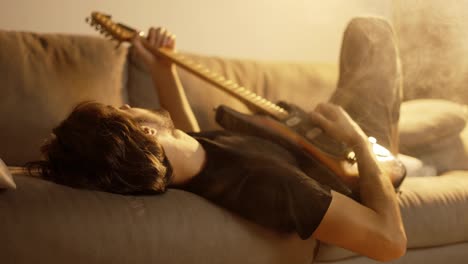 Man-playing-on-guitar-while-lying-on-couch-in-smoky-room