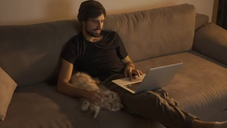 Hombre-Feliz-Trabajando-En-Una-Computadora-Portátil-En-Casa-Mientras-Se-Sienta-En-El-Sofá-Y-Acaricia-A-Un-Gato