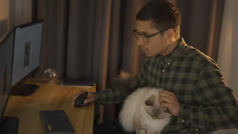 Junger-Mann,-Der-Vom-Heimbüro-Aus-Den-Online-Handel-Auf-Dem-Bildschirm-Betrachtet,-Sitzt-Mit-Einer-Süßen-Weißen,-Flauschigen-Katze