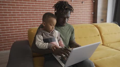 Joven-Padre-Afroamericano-Enseñando-A-Un-Niño-Pequeño-Enfocado-A-Usar-Una-Computadora-Portátil