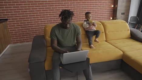 Busy-young-african-father-doing-freelance-work-on-laptop-and-babysitting-young-child-at-home