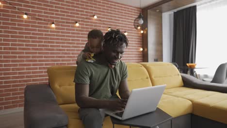 Smiling-african-father-doing-freelance-work-on-laptop-while-his-son-disturbing-him-from-the-back