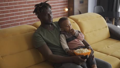 African-American-father-spending-time-with-small-son-sitting-on-couch-eating-popcorn-watching-TV