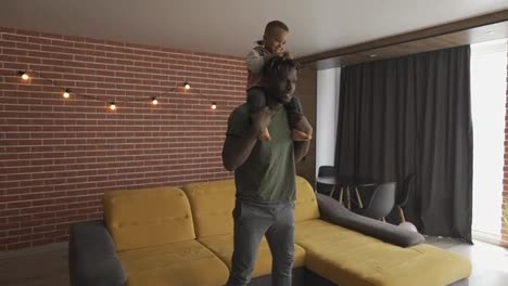Young-dad-does-squats-in-cozy-living-room,-his-toddler-son-sits-on-shoulders