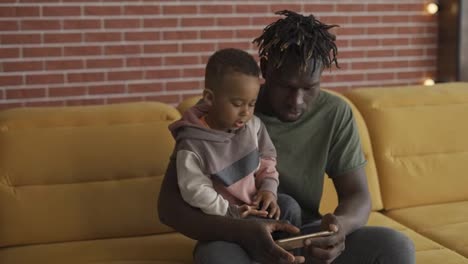 Portrait-of-african-american-father-sitting-with-son-on-sofa-using-smartphone
