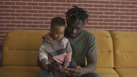 Portrait-of-african-american-father-sitting-with-son-on-sofa-watching-cartoon-on-smartphone