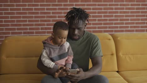 African-american-father-sitting-with-son-on-sofa-using-smartphone
