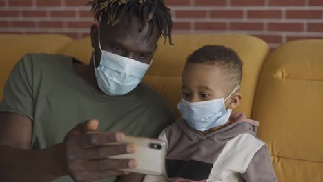 Father-and-son-are-sitting-on-a-sofa-in-medical-face-masks,-watching-video-on-smartphone