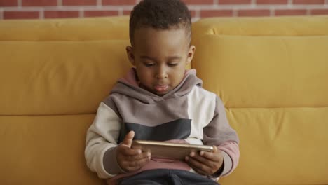 Afrikanischer-Kleiner-Süßer-Junge-Sitzt-Auf-Dem-Sofa-Und-Schaut-Sich-Mit-Dem-Smartphone-Cartoons-An,-Die-Spaß-Haben