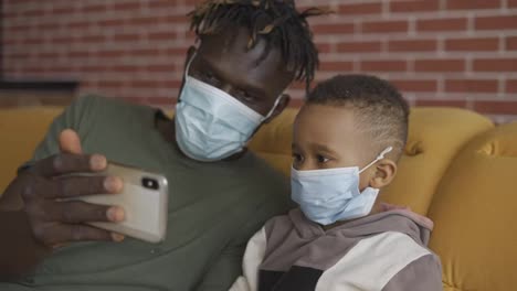 Father-and-son-are-sitting-on-a-sofa-in-medical-face-masks,-watching-video-on-smartphone,-close-up
