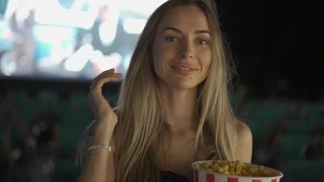 Blonde-woman-standing-with-bucket-of-popcorn-at-the-cinema,-smiling-to-the-camera