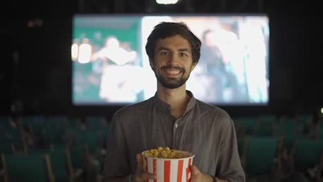 A-bearded-guy-standing-with-bucket-of-popcorn-at-the-cinema,-smiling-to-the-camera