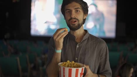 A-bearded-guy-eats-popcorn-at-the-summer-cinema,-smiling-to-the-camera