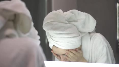 A-woman-with-towel-on-head-washes-her-face-with-water,-looking-at-the-mirror