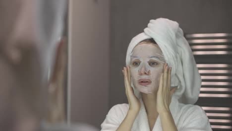 Woman-in-bathrobe-and-towel-with-white-mask-for-moisturizing-on-face,-looking-at-the-mirror